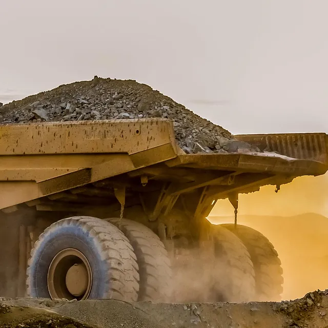 Stor dumptruck med last av stein kjører i støvete omgivelser ved solnedgang.