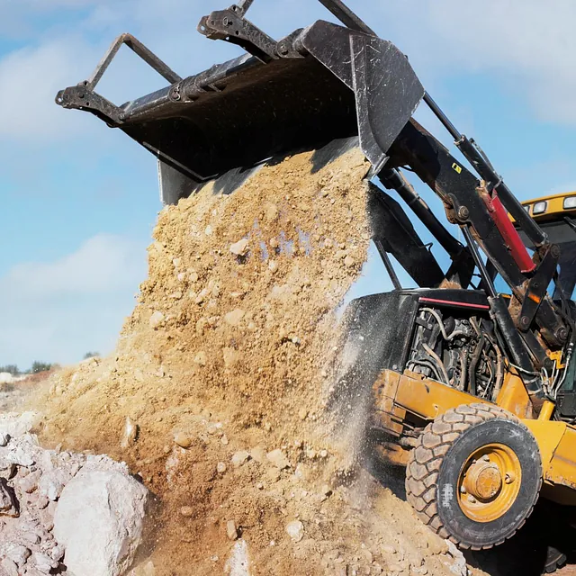 Gravemaskin tømmer sand og stein fra skuffen på byggeplass mot blå himmel.