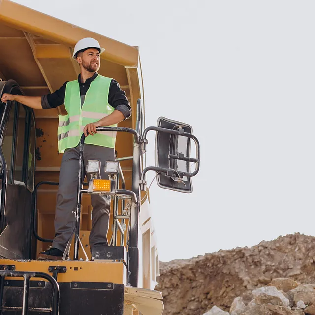 Byggearbeider i refleksvest og hjelm står på maskinplattform og ser utover byggeplass.
